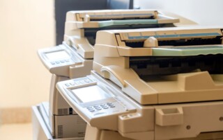 A row of professional copiers with document feeders and digital displays, set against a soft beige wall, indicative of a busy office setting where document duplication is frequently performed.
