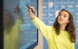 A young student in a bright yellow sweatshirt interacts with a touchscreen, writing or selecting options on a digital interface in a classroom setting. The background features a periodic table design, showcasing the use of interactive displays for educational purposes.