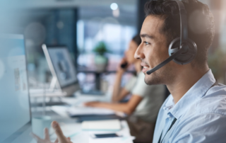 A focused male customer support agent wearing a headset with a microphone, providing IT services. He is engaging with a client in a bright, modern office environment, with blurred colleagues in the background also attending to customer queries