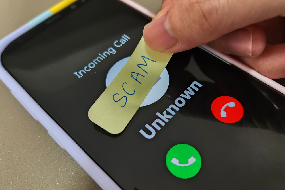A person's finger is placing a yellow sticky note with the word "SCAM" written on it over the screen of a smartphone, which displays an incoming call labeled "Unknown". This illustrates the concept of call screening, relevant to the STIR/SHAKEN initiative, which aims to identify and label suspicious calls.