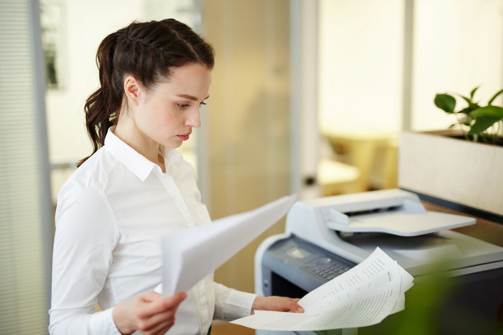 A businesswoman stands next to one of her office's copiers reviewing printed pieces of paper