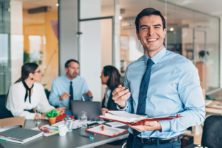 A smiling business professional in a blue shirt holding a notepad, standing in a modern office with colleagues discussing in the background. Managed Services Provider meeting in progress.