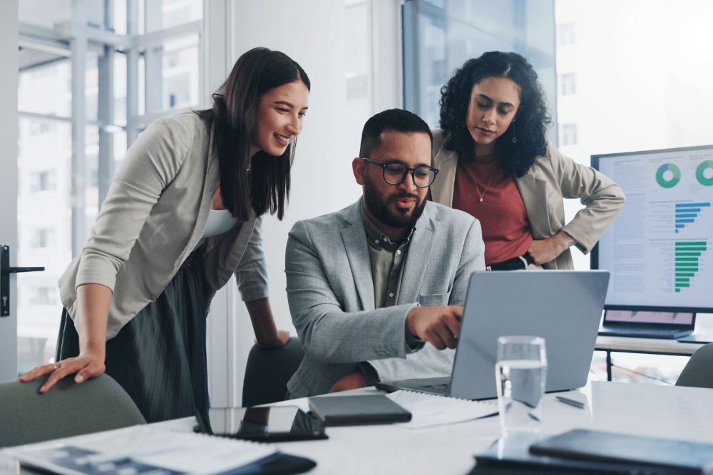 A group of young professionals stand around a laptop with a screen of charts and graphs in the background signifying Managed IT Services