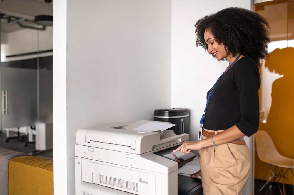 A young professional African American woman stands at an office laser printer as she interacts with the interface.