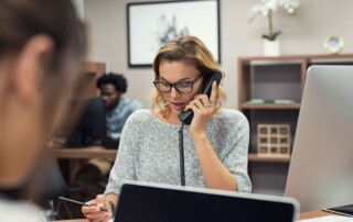 Close up of a woman wearing glasses at a desk in an office holding a pencil and speaking on a phone with other workers in front of her and behind her signifying VoIP phone systems.